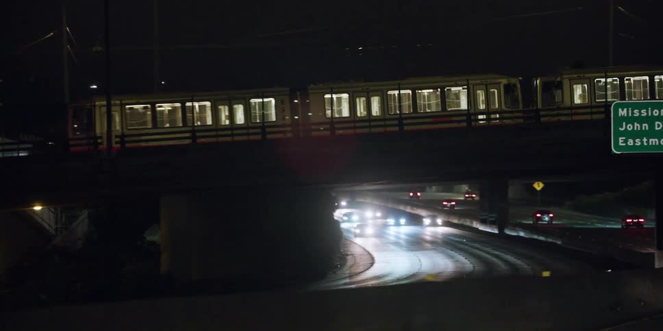 Automobiles are traveling on a freeway at night as a train passes on an overpass above the road