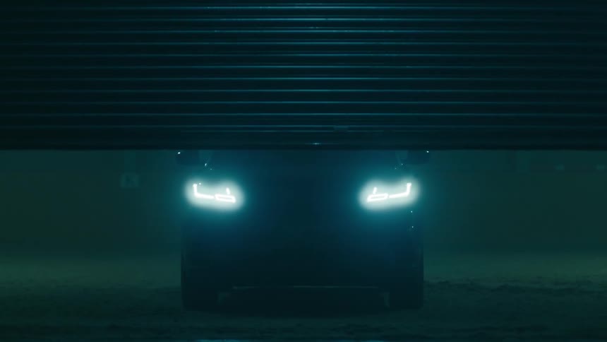 A garage door goes up and an automobile with the headlights on is sitting in the darkness