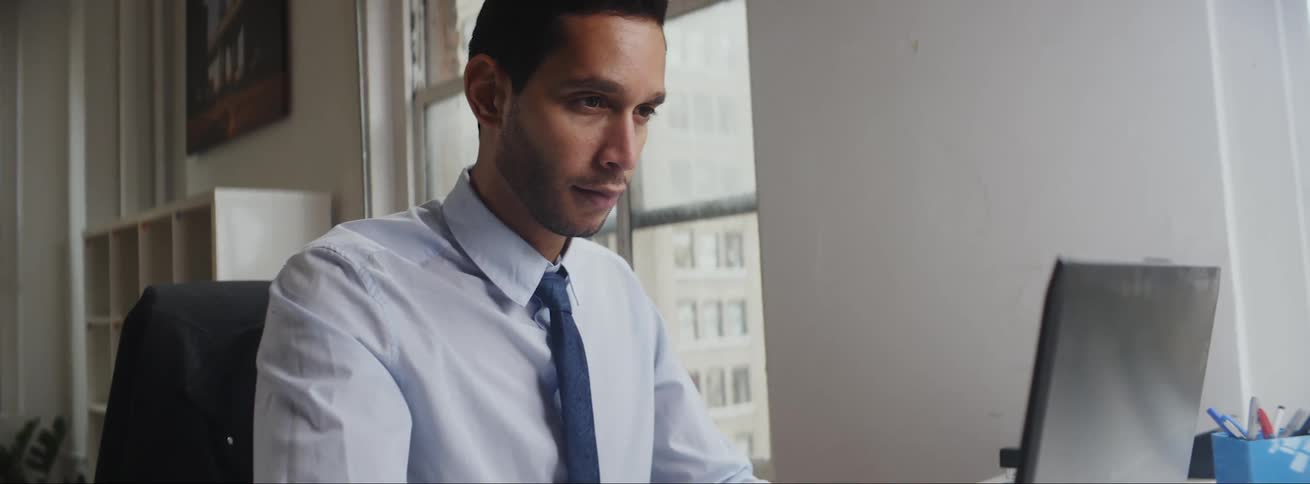 Businessman with dark blue tie and blue shirt sits at desk using laptop in the office
