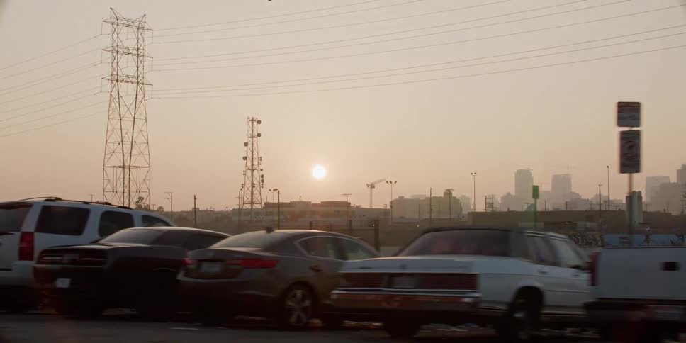 Automobiles are parked near utility towers and a bright sun is setting in the distance