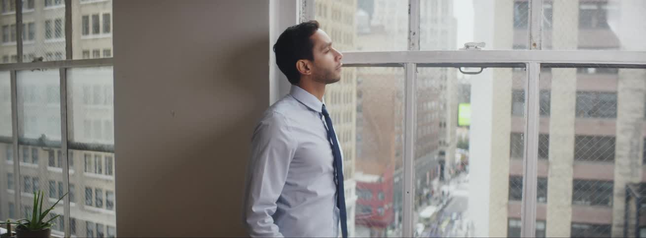 Businessman with shirt and tie looks out office windows overlooking city center