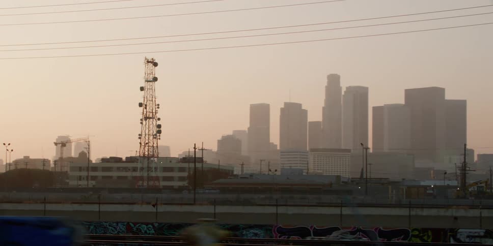 A utility tower stands near town with tall city buildings in the distance as people walk on the roadside