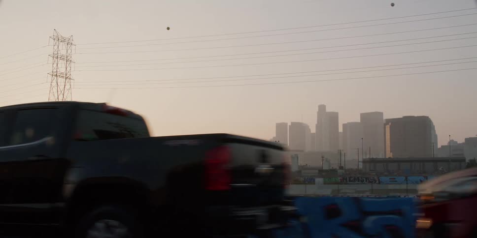 Cars and trucks are parked and there are utility towers and a city skyline in the distance with a bright sun in the sky