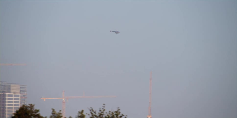 A helicopter flies high in the sky above trees and buildings