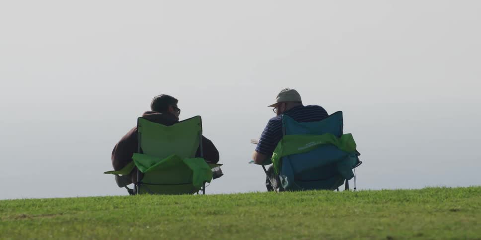 Two men sit in chairs on the shore and talk
