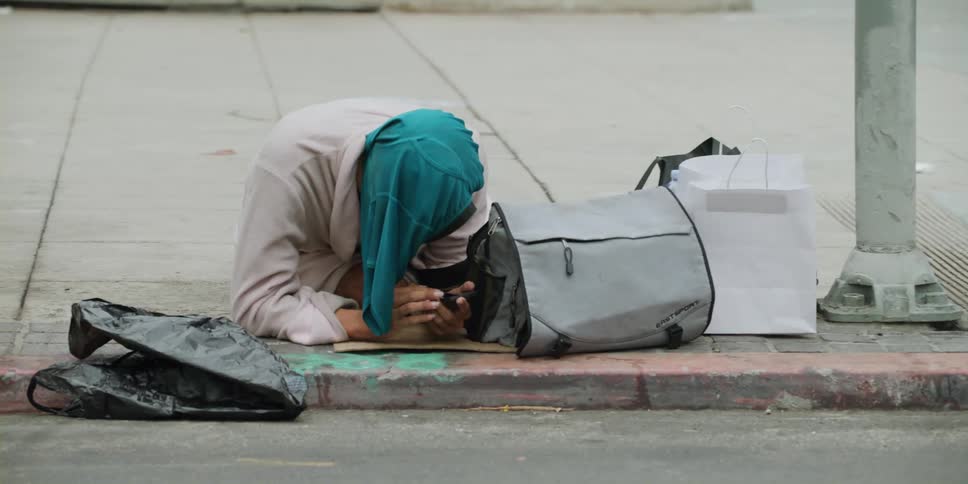 A homeless woman kneeling on a sidewalk near the curb with a shirt over her head tapping on a phone