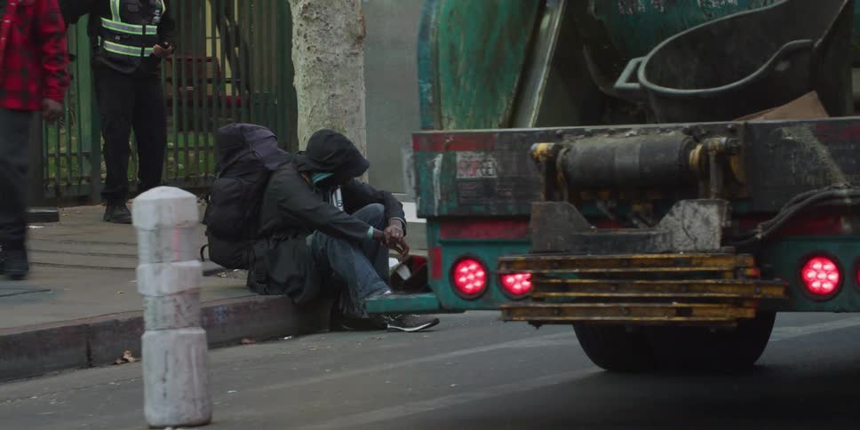 A man wearing a backpack sitting on the curb near a garbage truck looking dejected