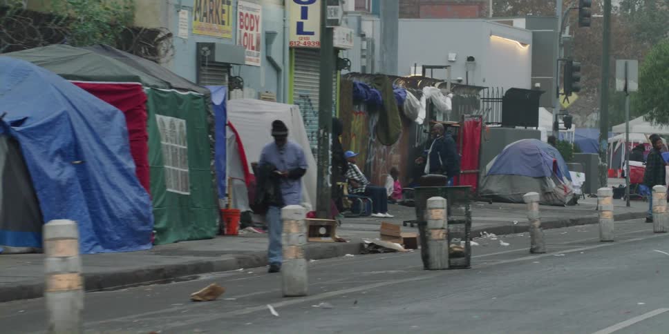 A car drives past people standing and walking on a sidewalk covered with tents