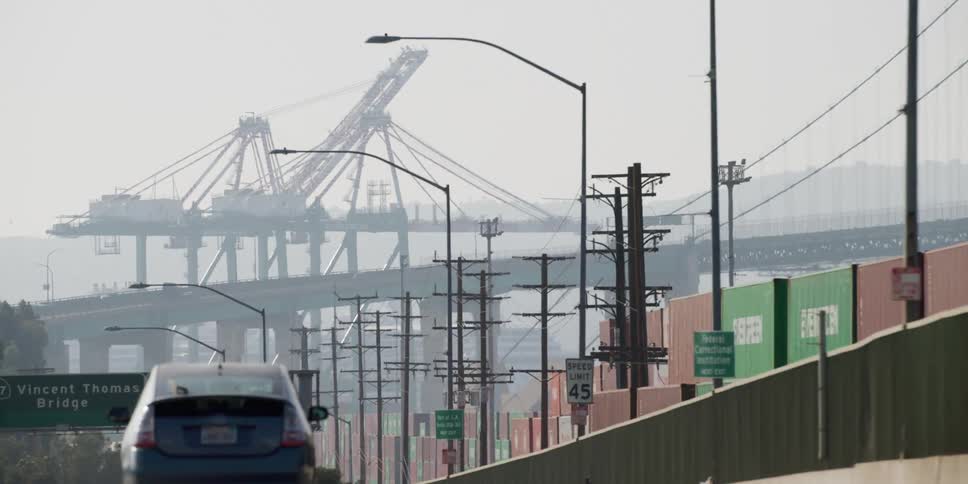 A vehicle travels on a highway bridge beside shipping containers and a large body of water