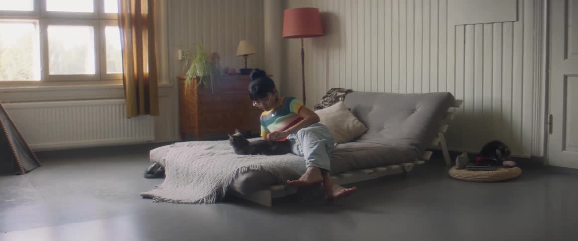 Girl with glasses stripped shirt and ripped jeans lies on bed petting gray cat in bedroom