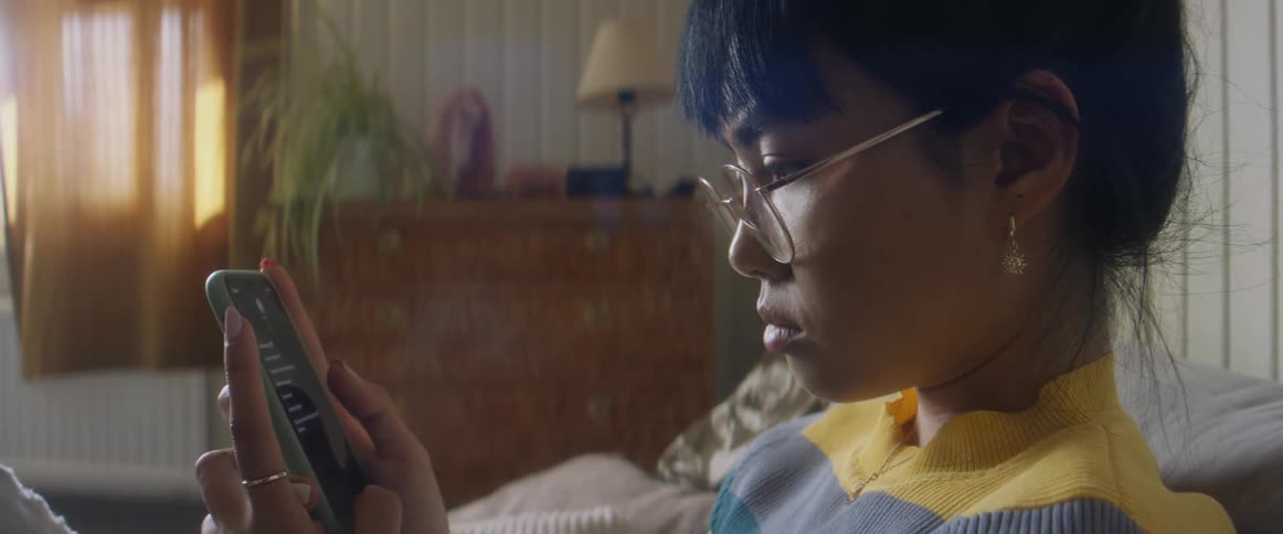 Girl with glasses earrings and stripped shirt sits on bed using smartphone in bedroom