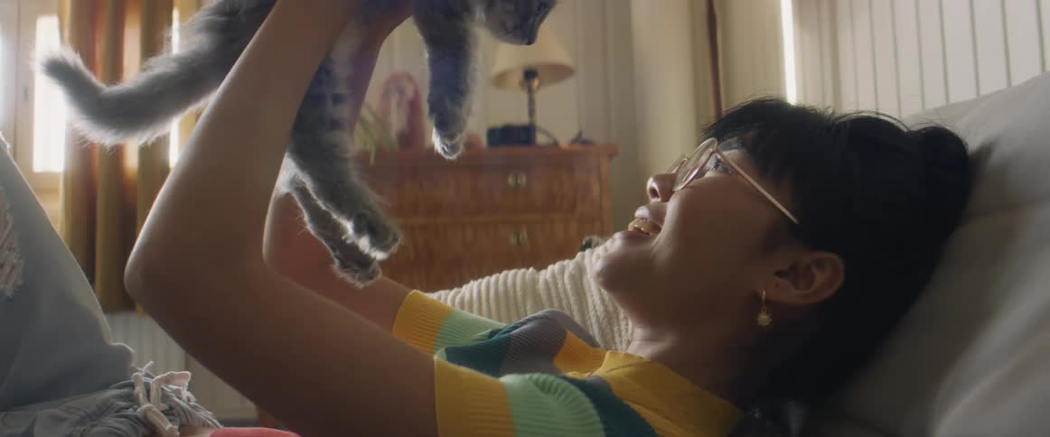 Girl with glasses earrings stripped shirt and jeans lies on bend petting gray cat in bedroom