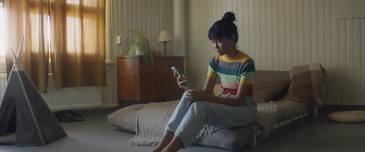 Girl with stripped tshirt and jeans sits on bed using smartphone in bedroom