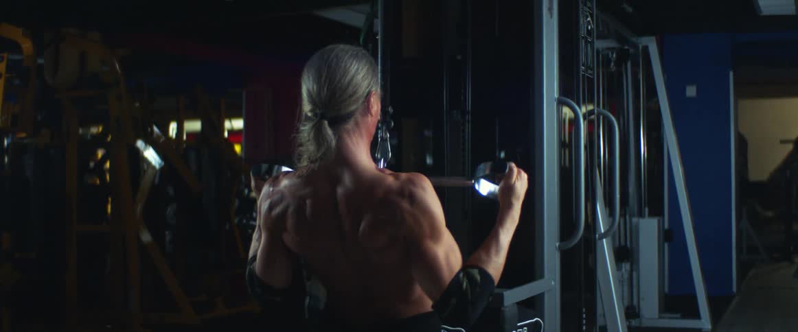 Shirtless blonde white man working out at an empty gym doing a back exercise at a gym equipment