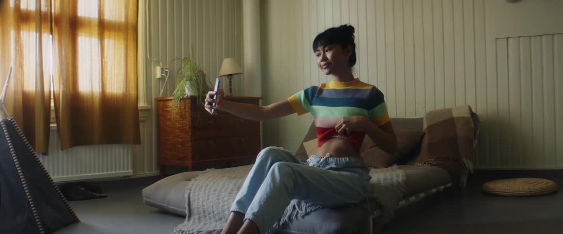 Girl with stripped tshirtand jeans sitting on bed takes selfie with smartphone in bedroom
