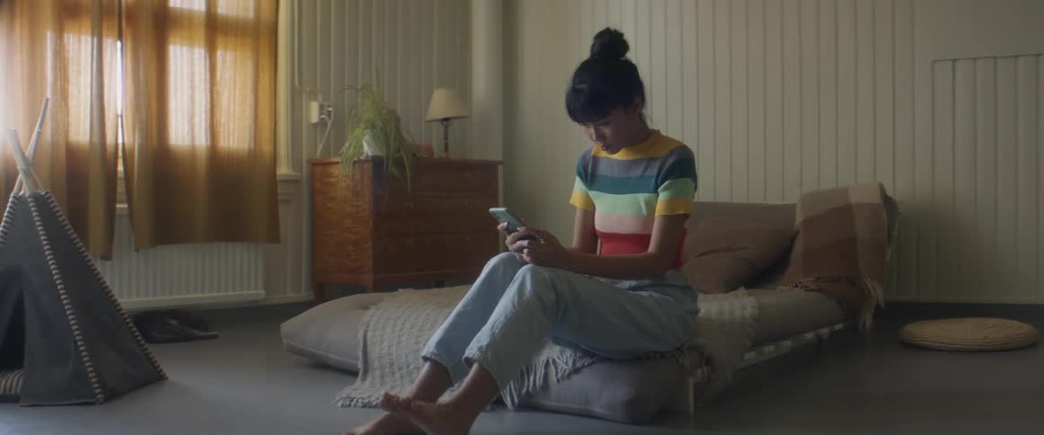 Woman with stripped shirt and ripped jeans sits on bed and uses smartphone in bedroom
