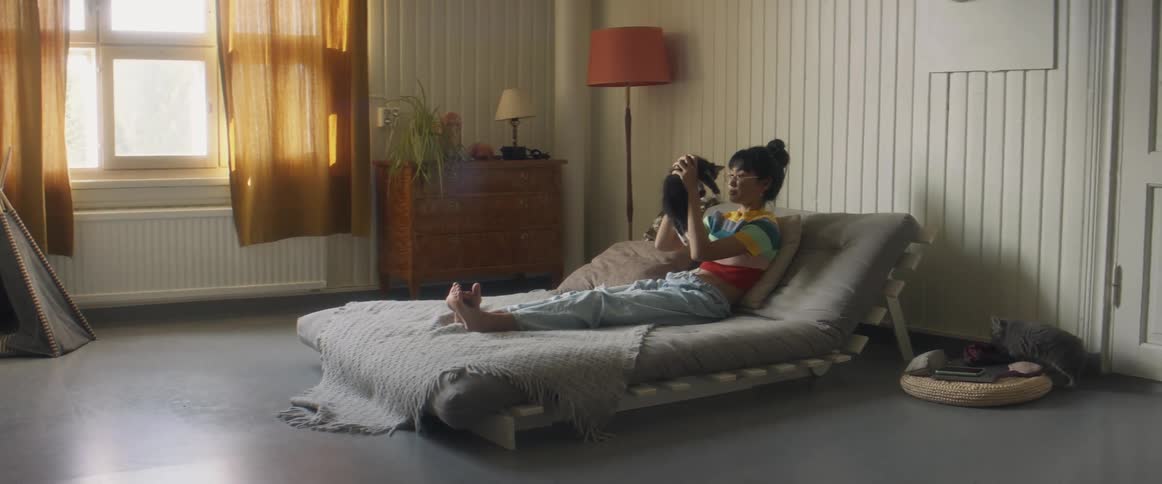 Girl with glasses stripped shirt and ripped jeans lying on bed pets and kisses gray cat in bedroom