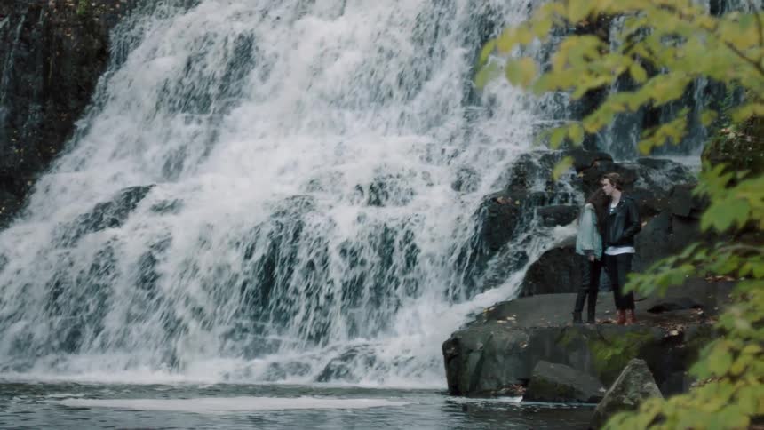 Two people are standing on rocks by a waterfall