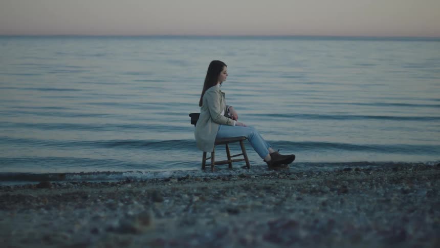 A woman sitting on a wooden chair as ocean waves gently roll in around her