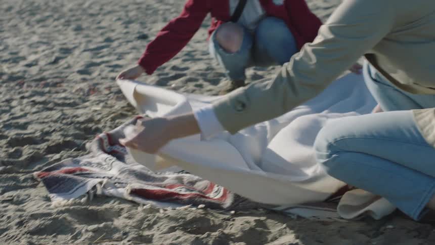 Two women laugh as they try to put a second blanket down on the beach sand on a windy day