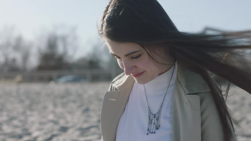 A woman on the beach looking down smiling softly while the wind blows her long hair