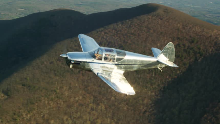 A pilot flies a fixed wing propeller plane over hills and peaks on a sunny day