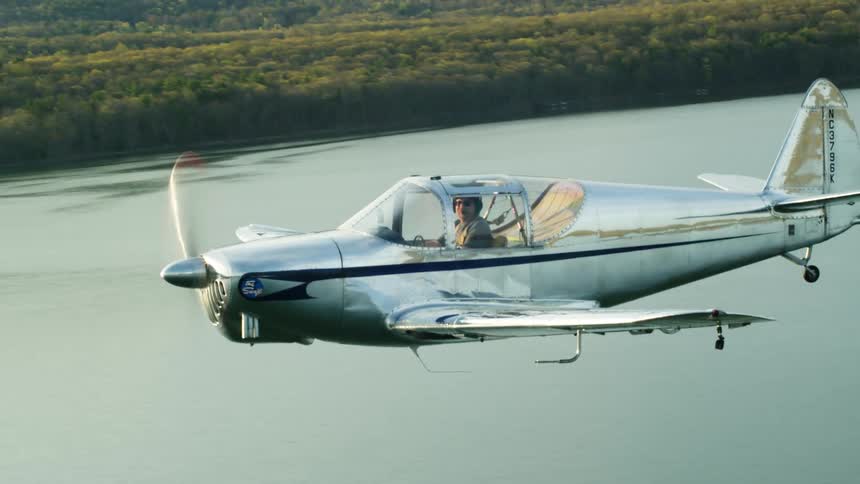 A man is flying a small airplane over a lake near a forest with mountains in the distance