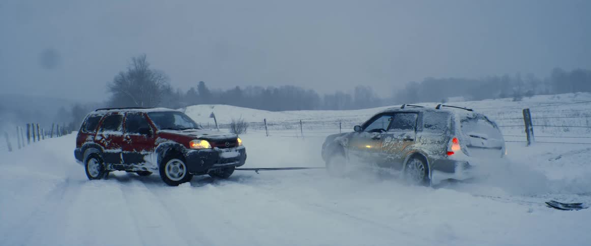 A vehicle uses a cable to pull another vehicle from a ditch on a snowy road in the countryside
