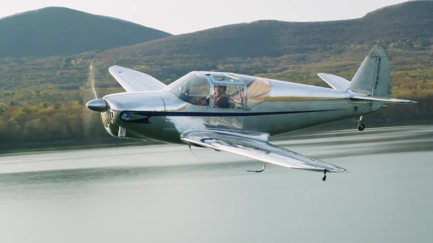 A pilot flies a propeller plane over the water and past trees with hills and mountains behind