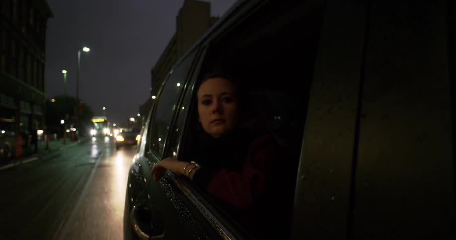 A woman partially leans out the window of a vehicle as it moves down a street at night