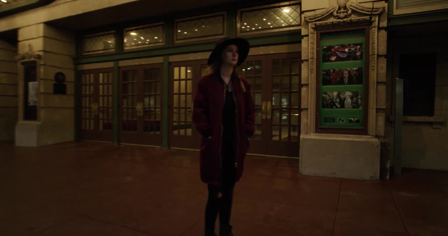 A woman wearing a big black hat stand in front of a theater in a city at night
