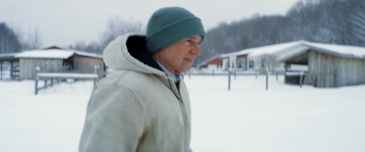 A man struggles a bit as he walks in the snow on his ranch pointing towards livestock and barn
