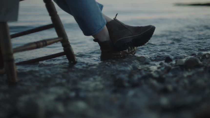 A woman sits on a chair on the shore as waves roll over her boots