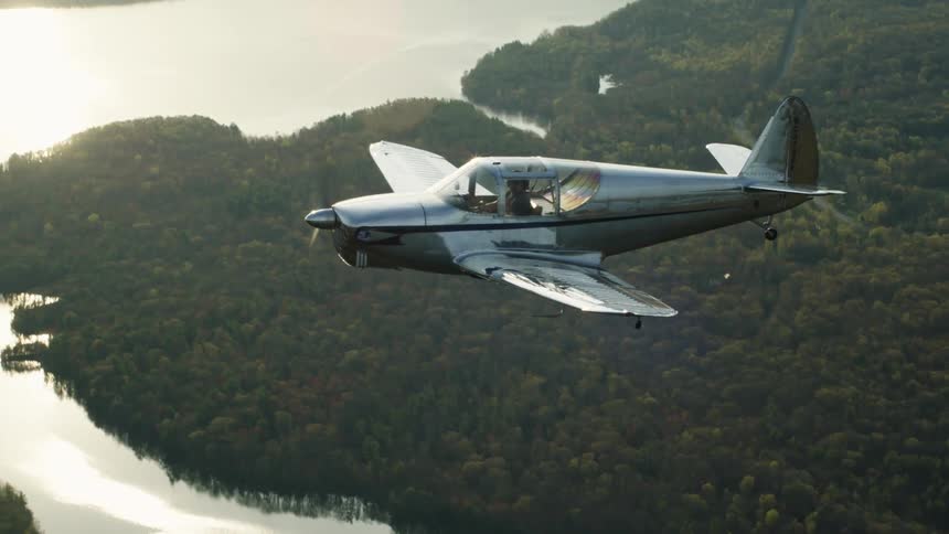 The sun is shining brightly upon a small airplane flying over a forest and lake