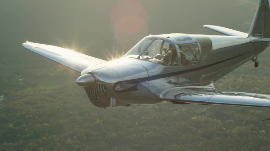 A man is flying a small airplane over a forest and lake with mountains in the distance