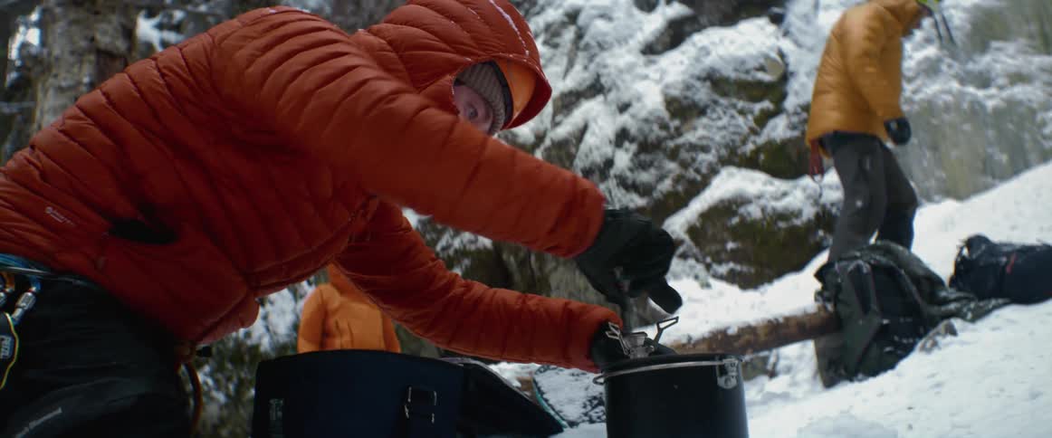 Man wearing a jacket adjusts a metal pot while another man walks up a snowy hill nearby
