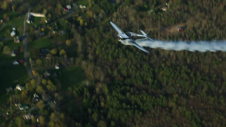 Metallic airplane trailing smoke performs roll over forest and nearby town