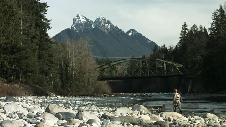 A fisherman wades through the river water carrying his pole near a bridge with a mountain on the horizon