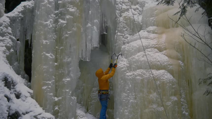 A climber is hammering a hook into the ice of a frozen waterfall preparing to climb it during the daytime