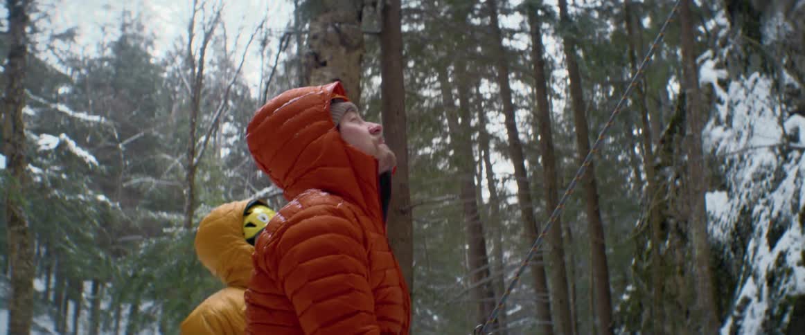 Man standing in a snow covered forest with another climber wears a climbing harness