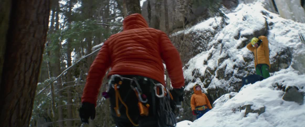 Two people stand on a snowy mountain as another person walks towards them with climbing gear on their pants