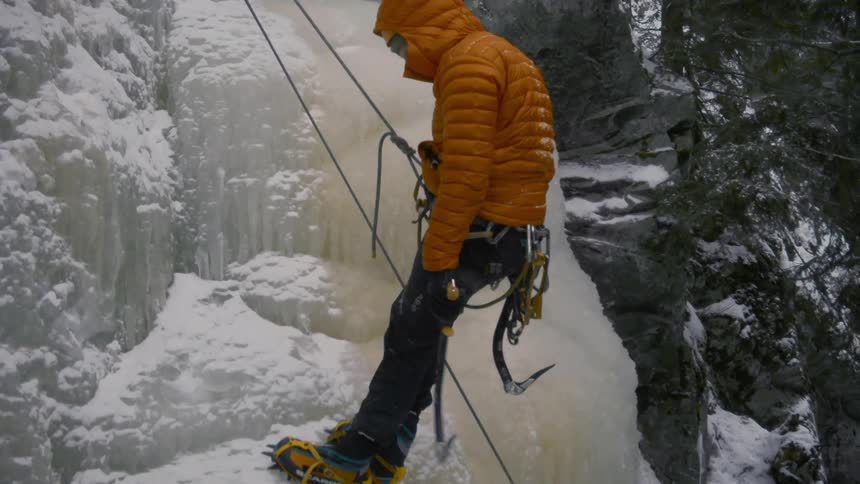 Man wearing climbing gear moves down ice covered mountain slope bordered by conifer forest