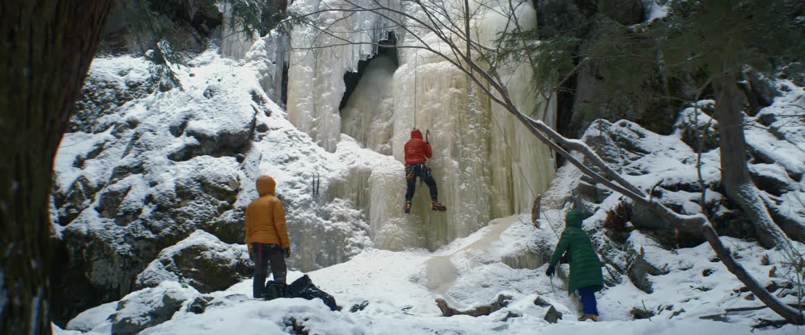 One person looks on and another helps a third climb ice wall