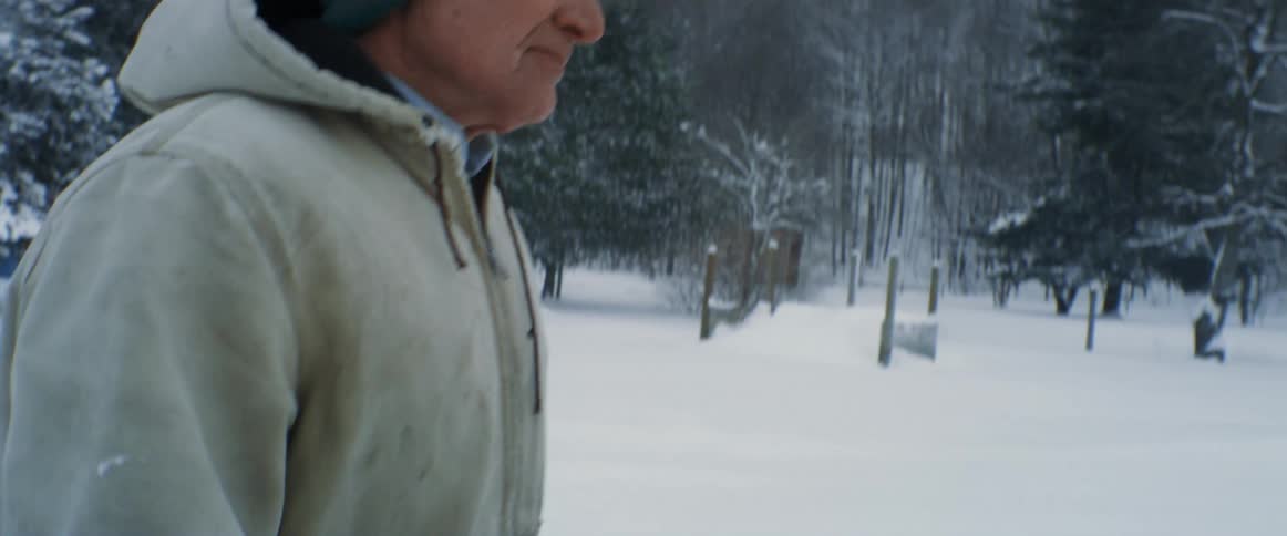 Old white man wearing a winter cap a coat and gloves walking alone in the snow near an arctic forest