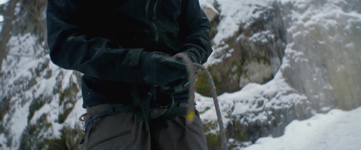 A rock climber ties a rope around his waist while standing in front of a snow covered ridge