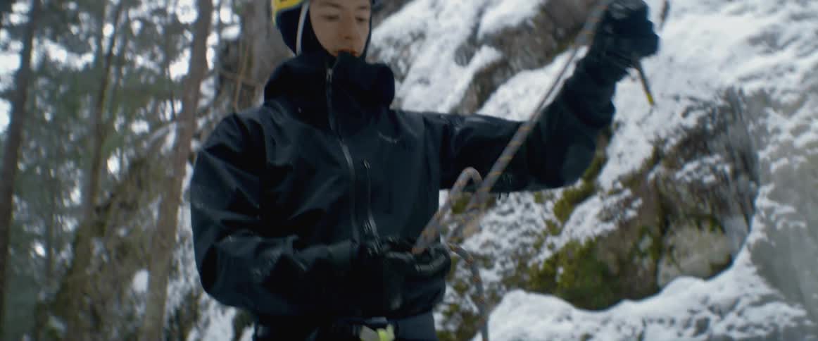 Mountaineer stands in conifer forest near snow covered mountain side and ties a knot in a rope