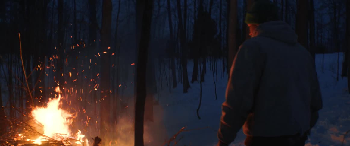 A man walks to a large fire of sticks in a wooded area