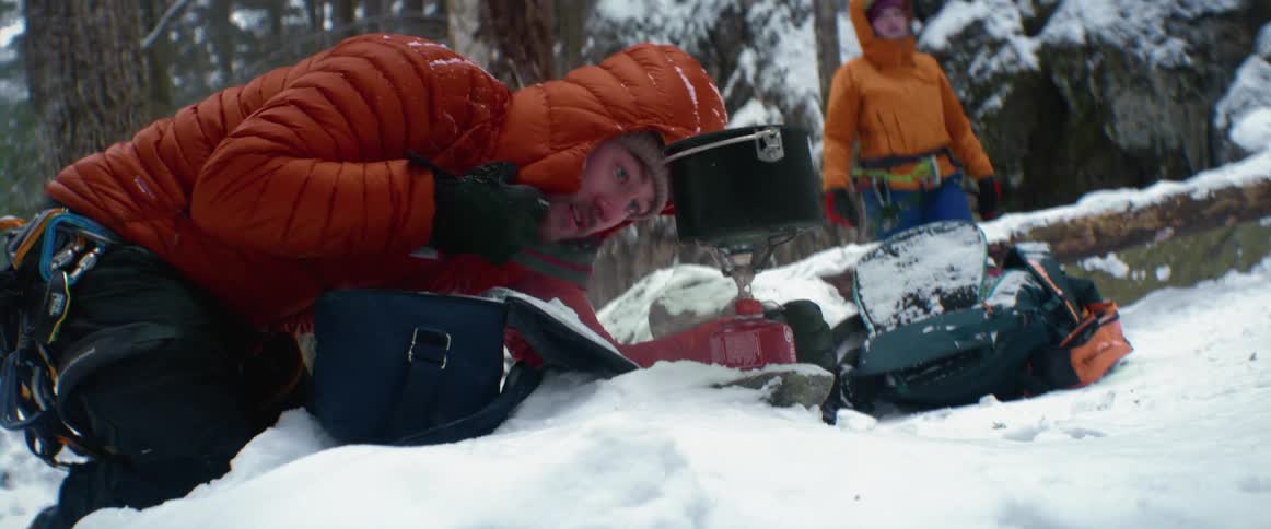 A man at the base of a snowy mountain tries to light a heater as a woman stands by and watches