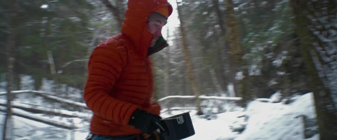 A person fills up a small pot from a creek in a snow covered woods area