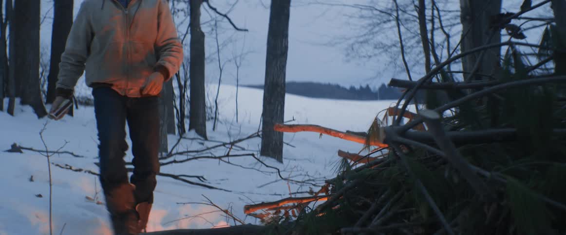 A man lights a large pile of sticks on fire in a snow covered woods area